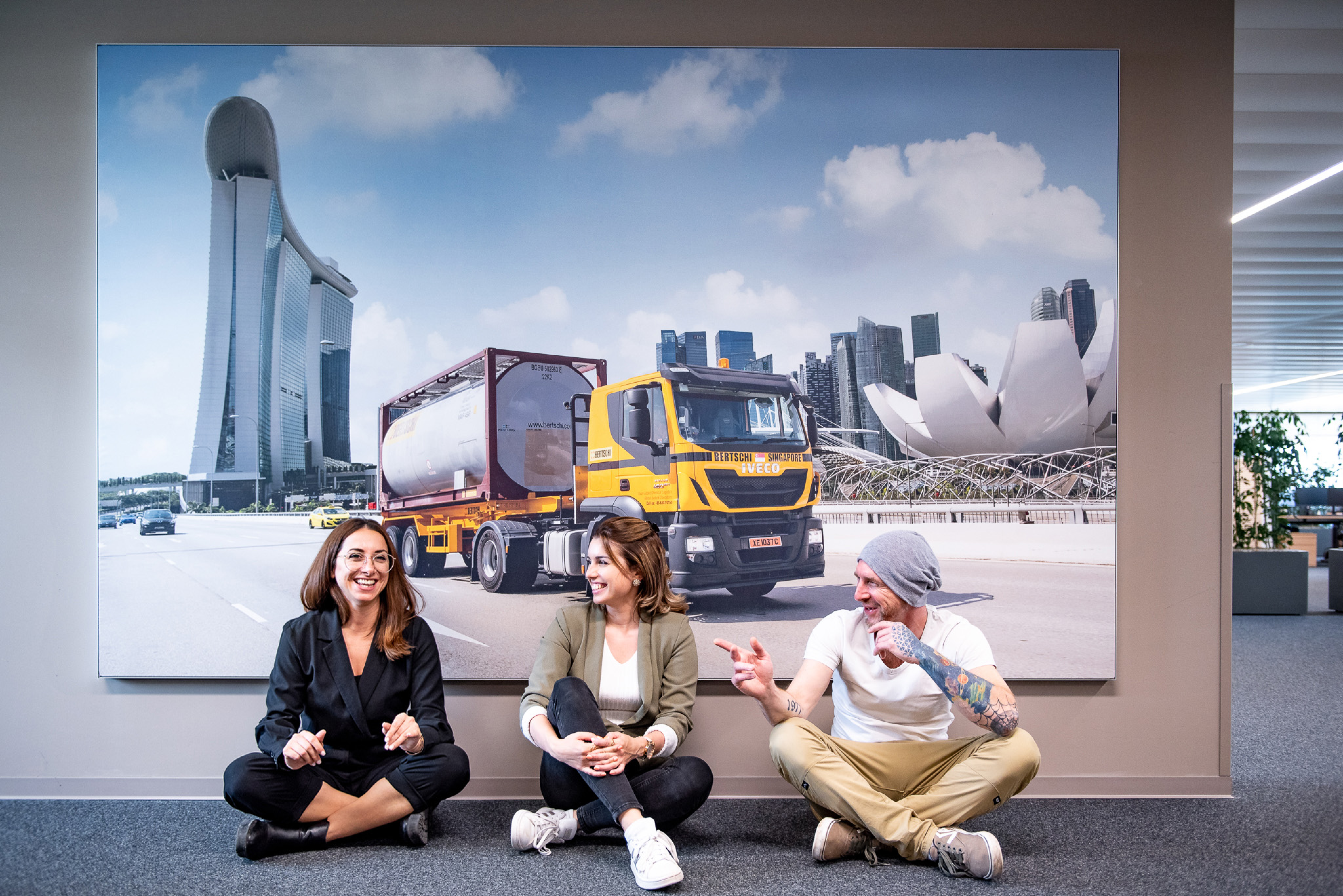 3 Employees sitting on the floor in front of a large picture of a Bertschi truck in Singapore